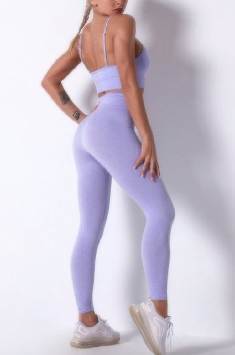 A woman modeling a matching lavender purple activewear set consisting of a sports bra and high-waisted leggings, paired with white sneakers, against a plain white studio background.