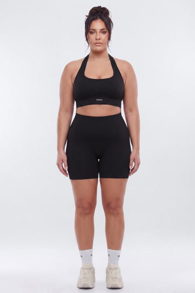 A woman modeling a matching black activewear set consisting of a halter crop top and high-waisted biker shorts, paired with white socks and beige sneakers, against a light gray studio background.