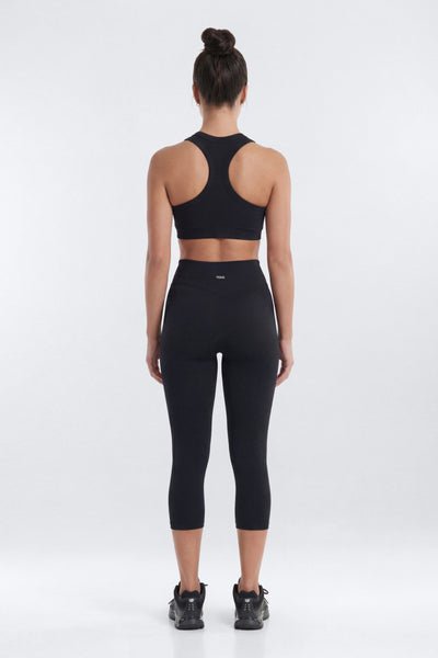 A woman viewed from behind wearing a black sports bra and matching black high-waisted capri leggings, standing against a light gray studio background.