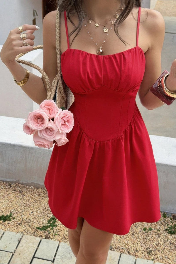 A woman wearing a bright red spaghetti-strap mini dress with a gathered bodice and flared skirt, holding a woven handbag with pink roses, in an outdoor setting.