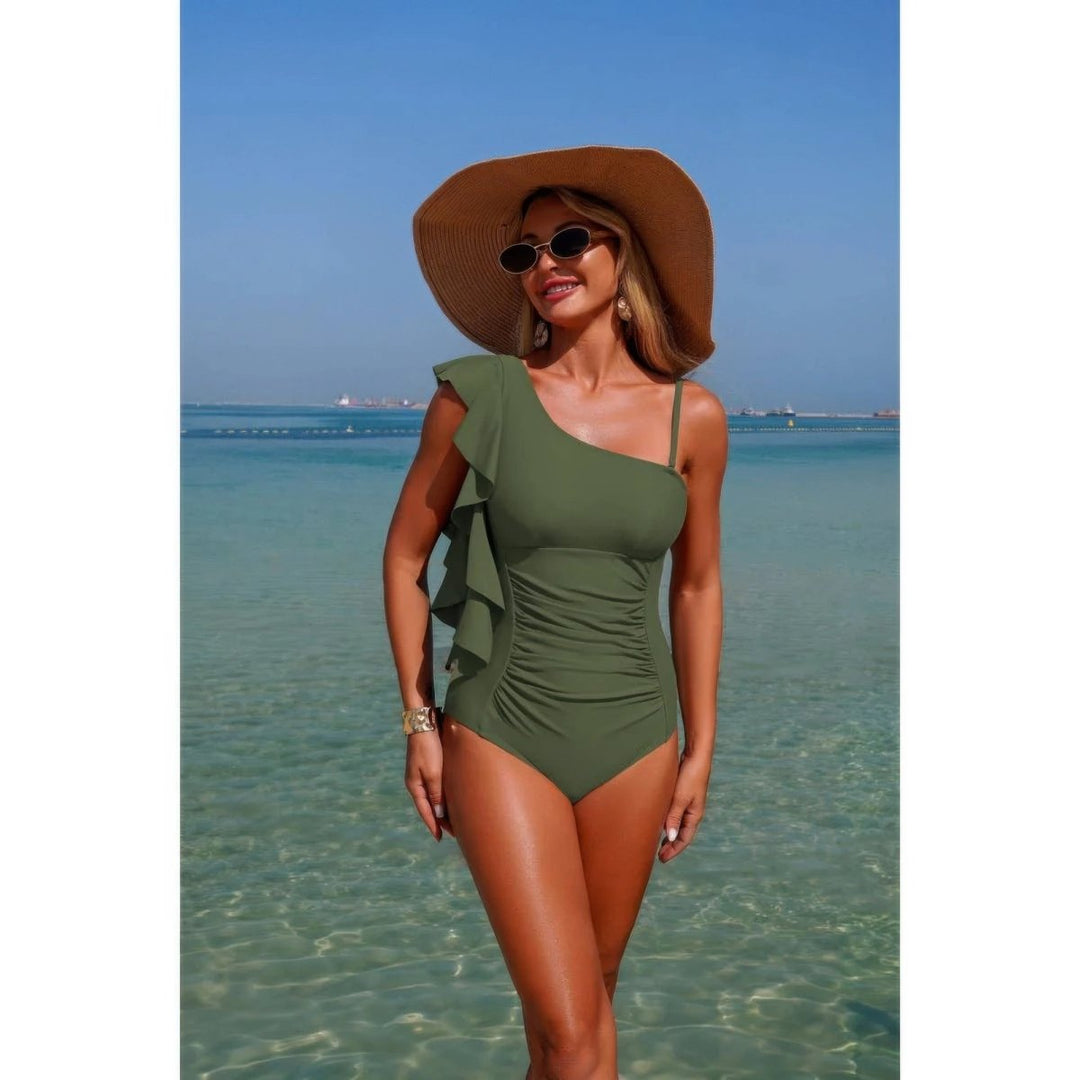 A woman wearing an olive green one-shoulder swimsuit with ruffle details, a wide-brimmed brown sun hat, and sunglasses, standing in shallow turquoise ocean water under a clear blue sky.