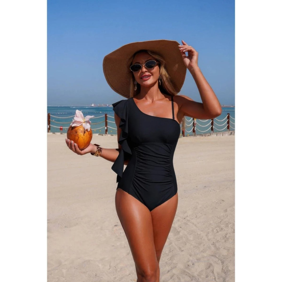A woman on a sandy beach wearing a black one-shoulder swimsuit with ruffle details, a large brown sun hat, and sunglasses, while holding a coconut drink.