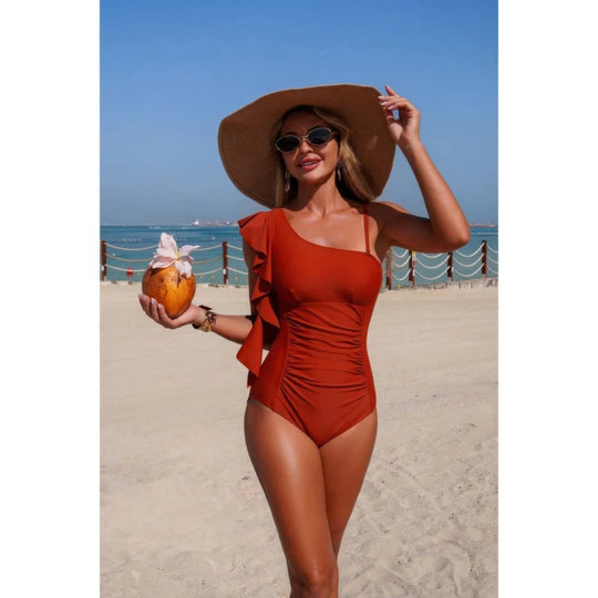 A woman stands on a sandy beach wearing a rust-orange one-shoulder ruched swimsuit, a large brown sun hat, and sunglasses, while holding a coconut drink.
