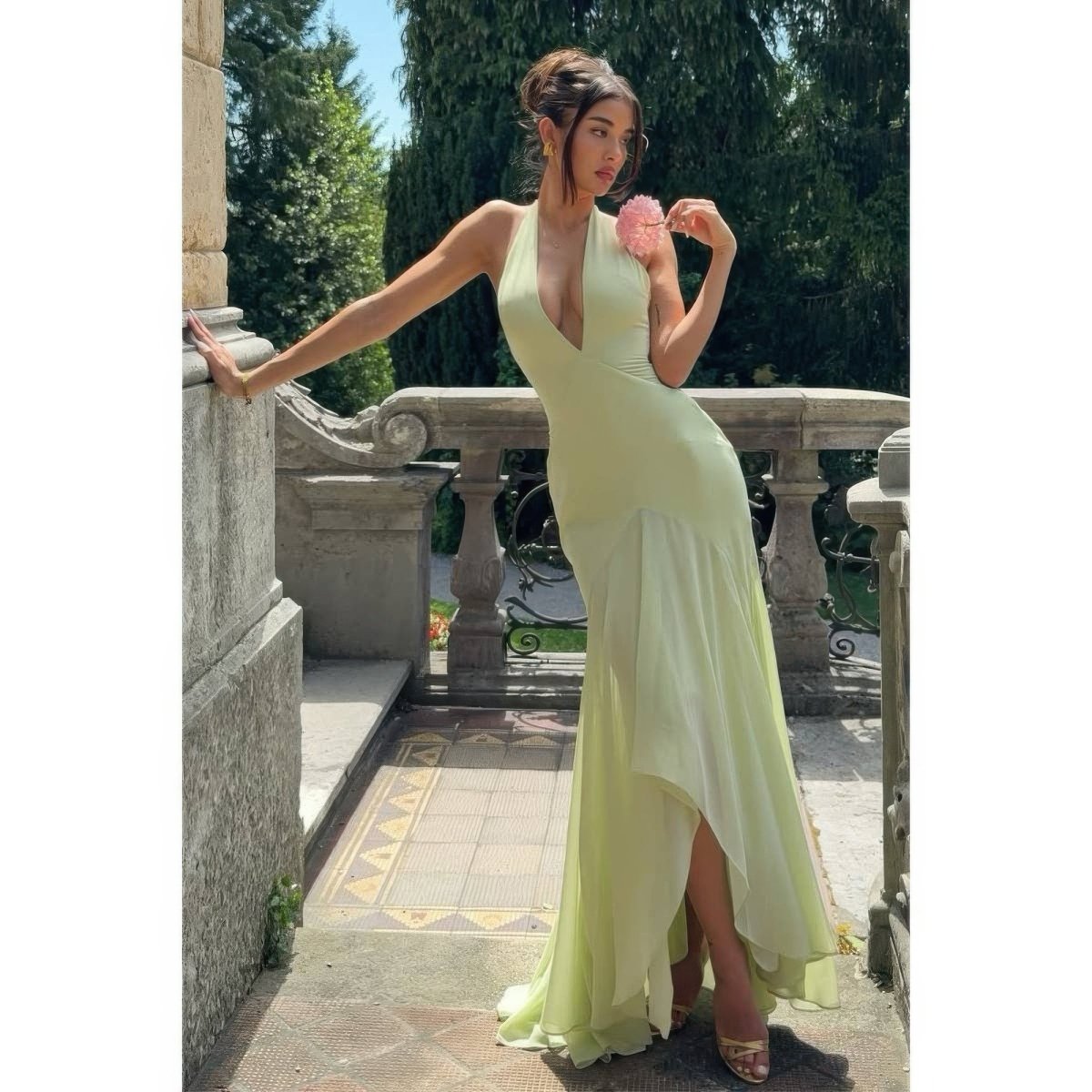 A woman wearing a pale yellow-green halter-neck maxi dress with a plunging V-neckline and a high-low skirt, posing on an outdoor stone terrace with trees in the background.