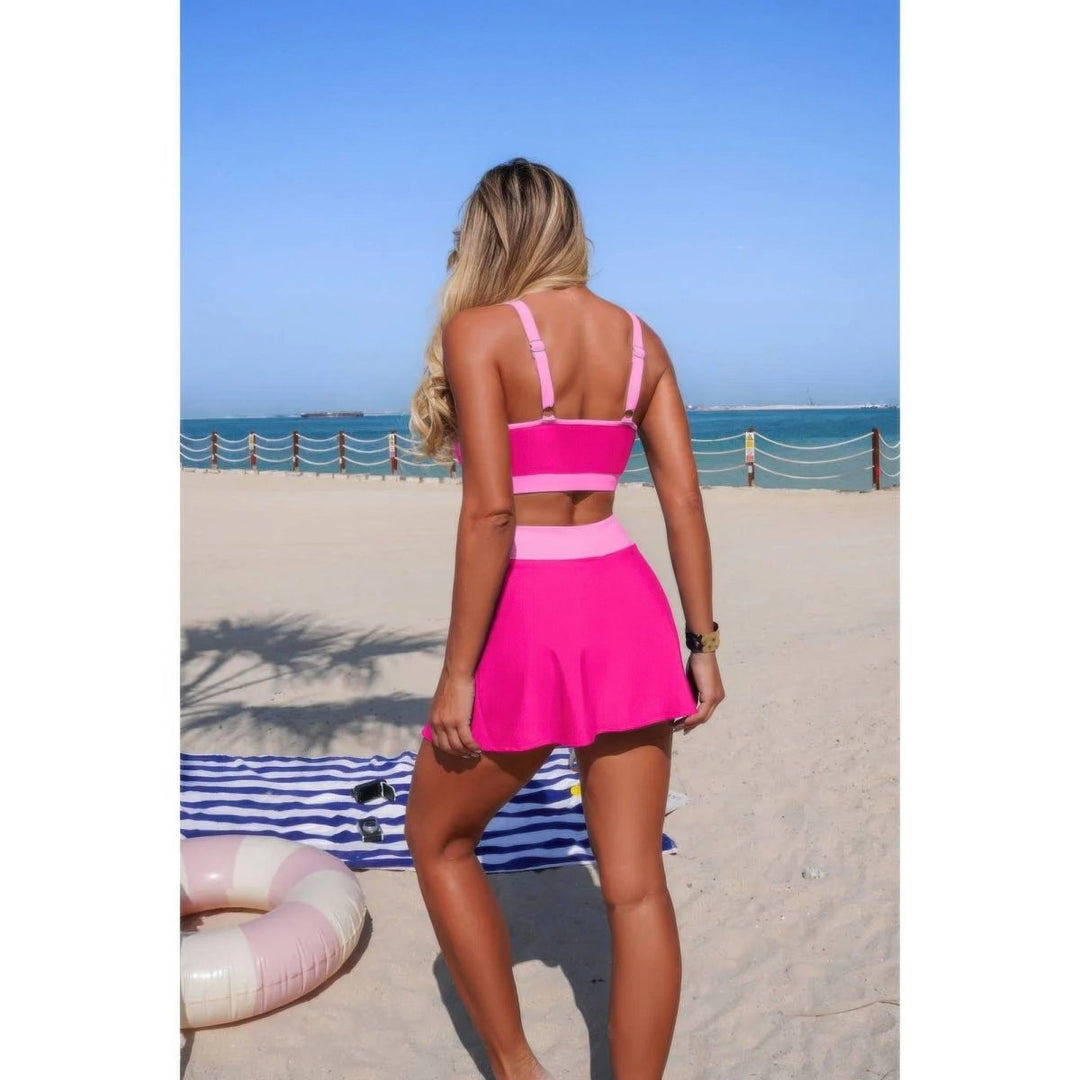 A woman stands on a sandy beach facing away from the camera, wearing a bright pink two-piece swimsuit with a strappy crop top and a flared mini skirt. The background shows the blue ocean, sky, and a striped beach towel.
