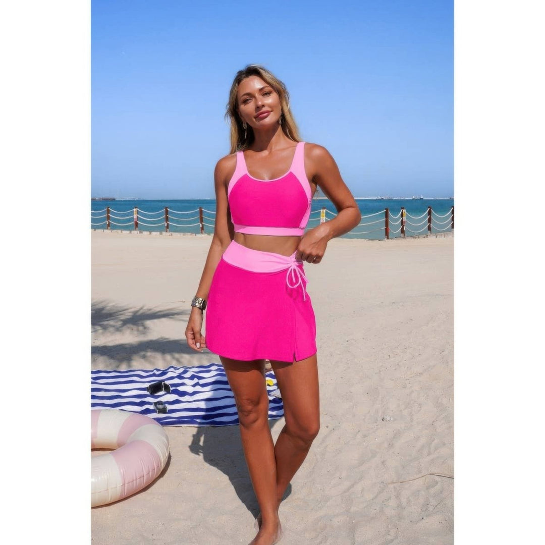 A woman modeling a bright pink two-piece swimsuit with a matching high-waisted tie-waist skirt on a sandy beach with the ocean and blue sky in the background.
