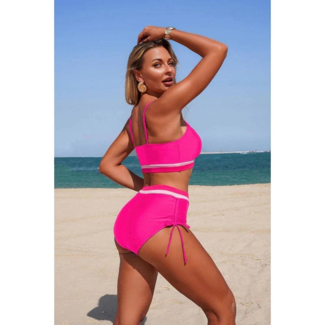 A woman posing on a sandy beach wearing a bright pink two-piece swimsuit with white stripes, with the ocean and blue sky in the background.