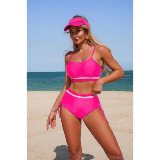 A woman modeling a bright pink two-piece bikini with white trim and a matching pink visor on a sandy beach with the ocean and blue sky in the background.