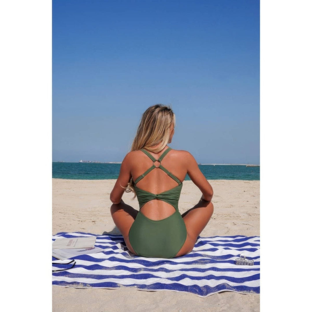 A woman sits on a blue and white striped beach towel on a sandy beach, viewed from behind, wearing an olive green one-piece swimsuit with crisscross back straps.