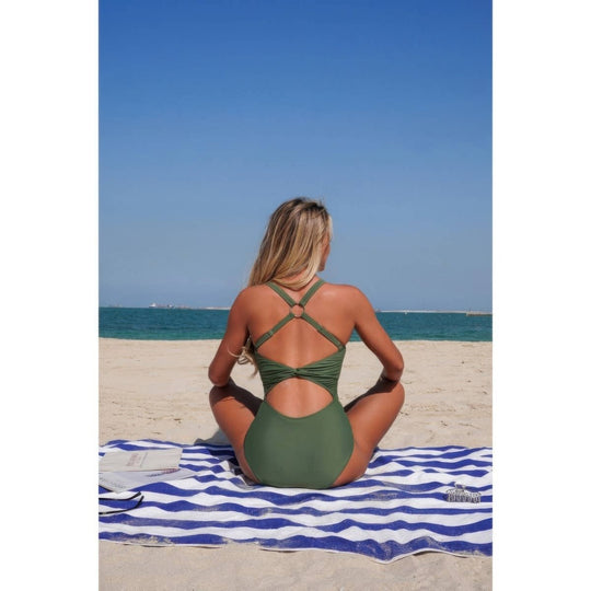 A woman sits on a blue and white striped beach towel on a sandy beach, viewed from behind, wearing an olive green one-piece swimsuit with crisscross back straps.