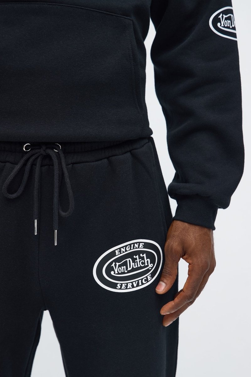 Close-up of a person wearing a black sweatshirt and matching sweatpants with white circular Von Dutch Service logos on the left thigh and sleeve.