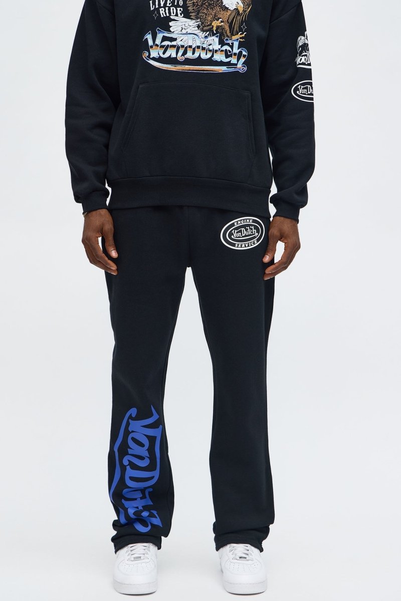 A person wearing a black hoodie and matching sweatpants with graphic prints, paired with white sneakers, photographed against a light gray studio background.