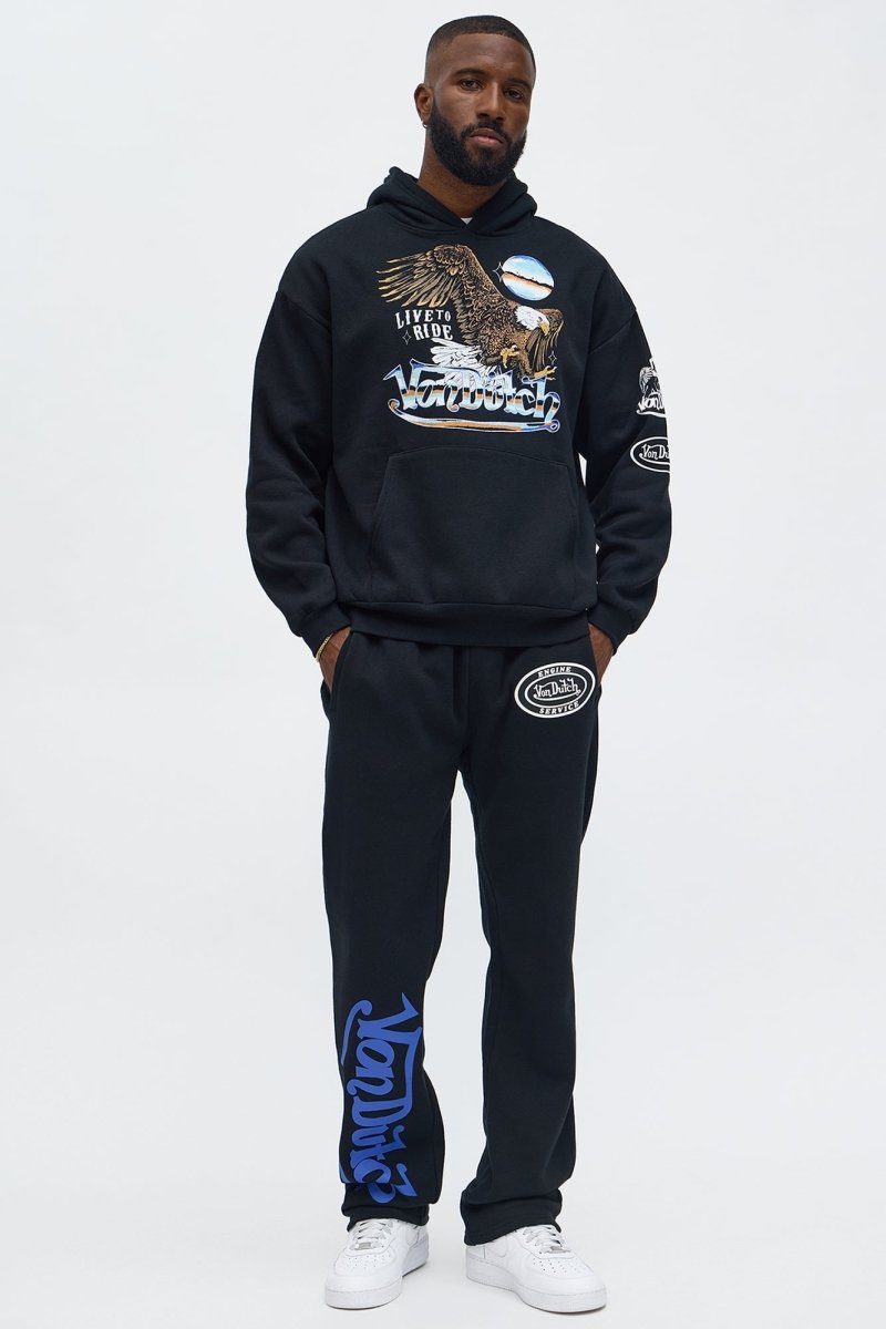 A man modeling a black streetwear tracksuit from the brand Natural, featuring a graphic hoodie with an eagle design. 